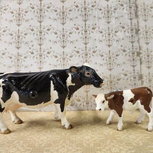May include: A black and white Holstein cow figurine standing next to a smaller brown and white calf figurine. Both animal figures are depicted in a standing position, featuring realistic coloring, textured surfaces, and detailed facial features.