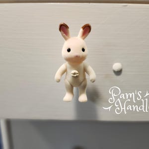 May include: A white rabbit figurine with brown ears and black eyes. The rabbit is wearing a small, white button on its chest. The figurine is on a white surface with the text "Pam's Handles" written in white.