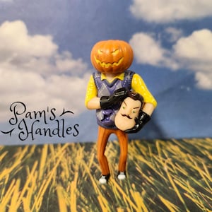 Può includere: Una statuina a tema Halloween con una testa di zucca intagliata e una figura che tiene una maschera. La figura indossa una camicia gialla, un gilet viola e pantaloni arancioni. Lo sfondo mostra un cielo blu con nuvole e un campo d'erba. Il testo "Pam's Handles" è nell'immagine.