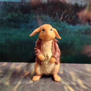 May include: A brown and tan rabbit figurine wearing a brown jacket. The rabbit has a white belly and a brown nose.