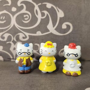 May include: Three white ceramic figurines of Hello Kitty characters. The character on the left is wearing a blue hat and brown vest. The character in the middle is wearing a yellow dress with a star. The character on the right is wearing a red headband and a white dress with a star.