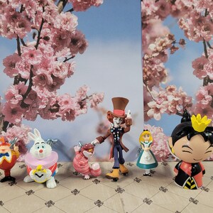Op de afbeelding: Een set van zes Alice in Wonderland-figuren, waaronder de Hoedenmaker, het Witte Konijn, de Cheshire Cat, Alice, de Koningin van harten en de tweeling Tweedledee en Tweedledum. De figuren zijn gemaakt van plastic en zijn geschilderd in felle kleuren.