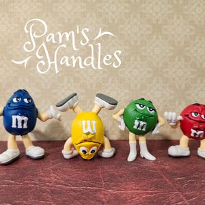 May include: Four colourful M&M's character figurines in blue, yellow, green, and red. Each figure has white gloves and shoes, with the letter "m" on the front. The text "Pam's Handles" is written in cursive above the figurines.