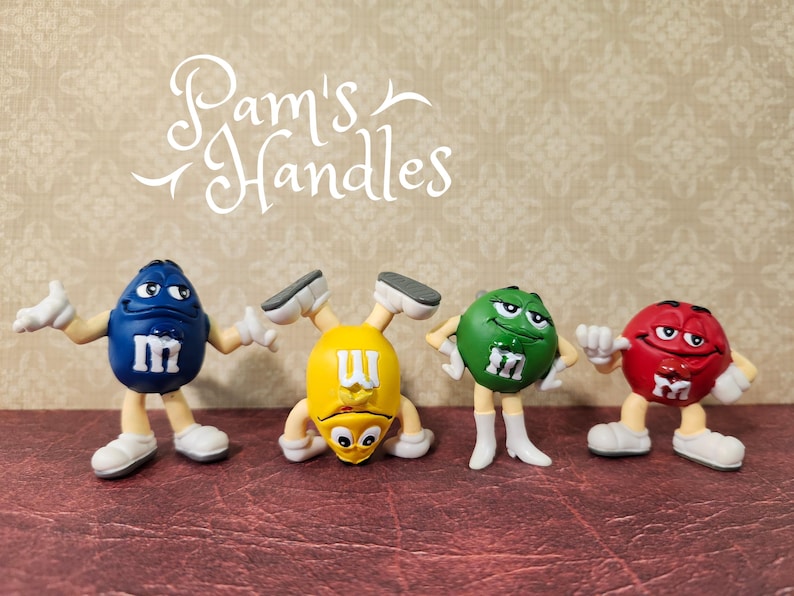Peut inclure: Quatre figurines color&eacute;es de personnages M&M's en bleu, jaune, vert et rouge. Chaque figurine a des gants et des chaussures blancs, avec la lettre "m" sur le devant. Le texte "Pam's Handles" est &eacute;crit en cursive au-dessus des figurines.