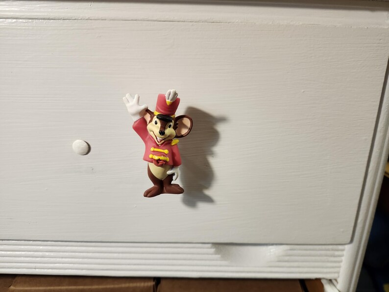 K&ouml;nnte beinhalten: Ein dekorativer Schubladengriff mit einer Zeichentrickmaus in roter und goldener Uniform, die mit einem wei&szlig;en Handschuh winkt. Die Maus hat braunes Fell, gro&szlig;e Ohren und einen schwarzen Hut. Der Griff ist an einer wei&szlig;en Kommode befestigt.