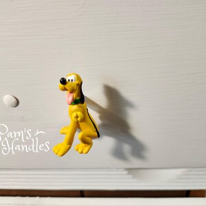 以下が含まれることがあります： 黒と緑のアクセントが付いた、黄色のプルート型のキャビネットノブ。ノブは白い引き出しに取り付けられています。白い丸いノブも表示されます。「Pam's Handles」というテキストが左下にあります。