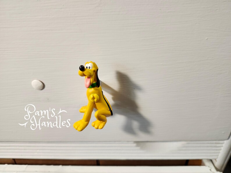 以下が含まれることがあります： 黒と緑のアクセントが付いた、黄色のプルート型のキャビネットノブ。ノブは白い引き出しに取り付けられています。白い丸いノブも表示されます。「Pam's Handles」というテキストが左下にあります。