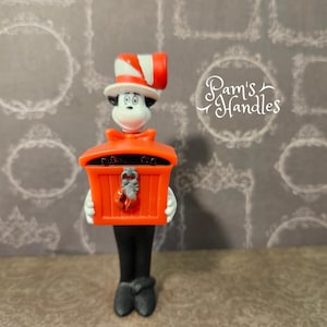 May include: A Cat in the Hat figurine holding an orange box. The figure wears a red and white striped hat, a red bow tie, and black trousers. The box has a silver clasp and a small red object.