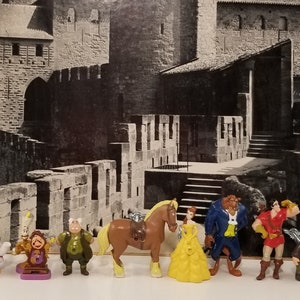 Könnte beinhalten: Ein Set aus neun Plastikfiguren aus dem Disney-Film Die Schöne und das Biest. Die Figuren beinhalten Belle, das Biest, Gaston, Lumière, Cogsworth, Mrs. Potts, Chip und das verzauberte Pferd. Die Figuren sind vor einem Schwarzweißfoto eines Schlosses platziert.