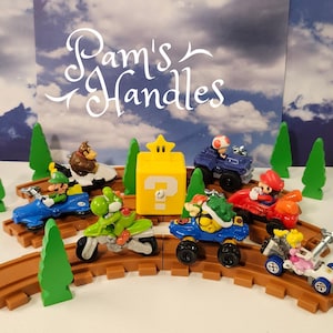 May include: A collection of Super Mario character figurines in various vehicles, including karts and a motorcycle, arranged on brown plastic track pieces with green tree cutouts and a yellow question mark block with a star on top.