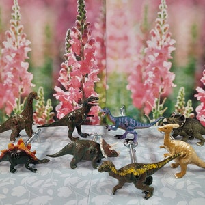 Może przedstawiać: Kolekcja dziewięciu kolorowych figurek dinozaurów, w tym zielono-brązowego Tyranozaura Rexa, fioletowego Welociraptora, brązowego Triceratopsa, brązowego Stegozaura i beżowego Parasaurolopha. Dinozaury są ułożone na białym tle z kwiatowym wzorem.