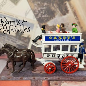 Könnte beinhalten: Eine Miniatur eines weißen Pferdewagens mit roten Rädern und dem Text "Oakey's Knife Polish". Der Wagen hat mehrere Miniaturpassagiere und wird von zwei braunen Pferden gezogen. Der Text "Pam's Handles" befindet sich oben links.