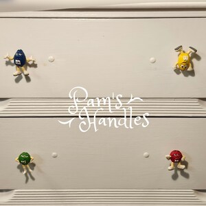 Peut inclure: Une commode blanche avec quatre boutons de tiroir repr&eacute;sentant des personnages M&M's color&eacute;s. Les boutons sont bleus, jaunes, verts et rouges. Le texte "Pam's Handles" est &eacute;crit en cursive au centre.