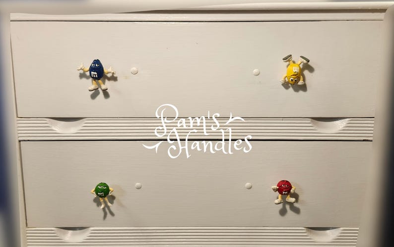 Peut inclure: Une commode blanche avec quatre boutons de tiroir repr&eacute;sentant des personnages M&M's color&eacute;s. Les boutons sont bleus, jaunes, verts et rouges. Le texte "Pam's Handles" est &eacute;crit en cursive au centre.