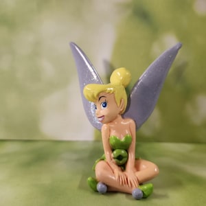 May include: A figurine of Tinkerbell, a character from Disney's Peter Pan. She has blonde hair, blue eyes, and wears a green dress. Her wings are a shimmering lavender color. She is seated with her legs crossed.