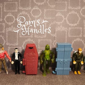 May include: A collection of vintage monster-themed action figures. The figures include a scarecrow, Dracula, a coffin, a swamp creature, a blue door, a man in a military uniform, and Frankenstein's monster. The background features a patterned wallpaper with the text "Pam's Handles".