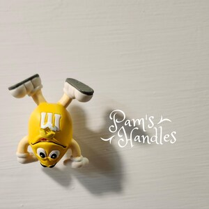 Peut inclure: Figurine jaune repr&eacute;sentant un personnage M&M's avec des chaussures et des bras blancs, et un visage noir et blanc. La figurine est &agrave; l'envers. Le texte "Pam's Handles" est en arri&egrave;re-plan.