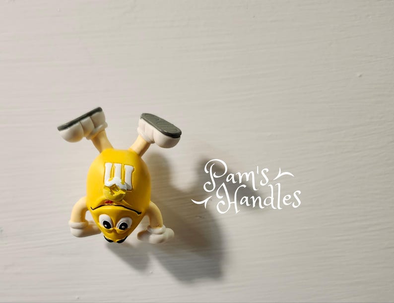 Peut inclure: Figurine jaune repr&eacute;sentant un personnage M&M's avec des chaussures et des bras blancs, et un visage noir et blanc. La figurine est &agrave; l'envers. Le texte "Pam's Handles" est en arri&egrave;re-plan.