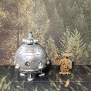 May include: A silver, round, spaceship with a dome-shaped top and three legs. A small, brown, alien figure with large eyes and a pointed finger is standing to the right of the spaceship. The background is a blurry image of a forest.