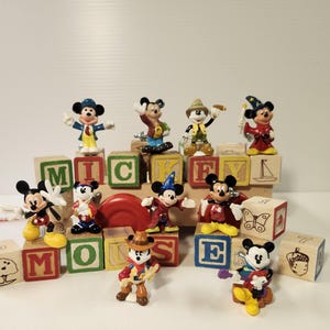 May include: A collection of ten small Mickey Mouse figurines in various costumes, including a wizard, cowboy, and explorer, arranged around and on top of several wooden alphabet blocks featuring letters and illustrations like a butterfly and acorn.