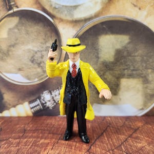 Op de afbeelding: Een gele trenchcoat met een zwart pak en een rode das, een zwarte fedora-hoed en een zwart pistool in de rechterhand. Het figuur is een plastic figuur.