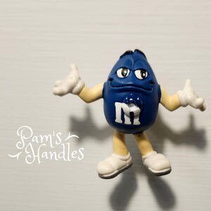 Peut inclure: Figurine de personnage de bonbon M&M's bleu avec des gants et des chaussures blancs. La figurine porte un "M" blanc sur le devant et est pos&eacute;e avec les bras tendus. Le fond est une surface gris clair.