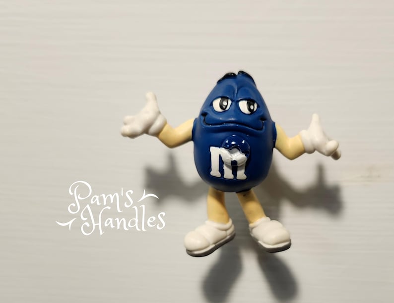 Peut inclure: Figurine de personnage de bonbon M&M's bleu avec des gants et des chaussures blancs. La figurine porte un "M" blanc sur le devant et est pos&eacute;e avec les bras tendus. Le fond est une surface gris clair.