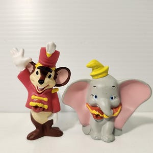 K&ouml;nnte beinhalten: Zwei Disney-Figuren: eine Maus in roter Uniform und ein grauer Elefant mit rosa Ohren. Die Maus winkt, und der Elefant tr&auml;gt einen gelben Hut. Beide Figuren haben detaillierte Merkmale.