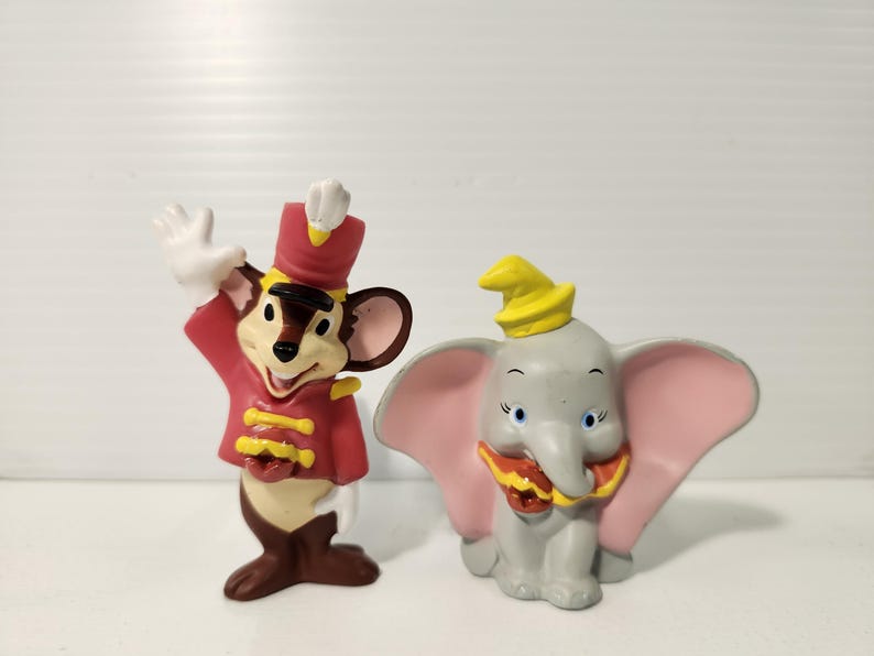 K&ouml;nnte beinhalten: Zwei Disney-Figuren: eine Maus in roter Uniform und ein grauer Elefant mit rosa Ohren. Die Maus winkt, und der Elefant tr&auml;gt einen gelben Hut. Beide Figuren haben detaillierte Merkmale.