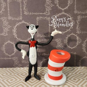 Puede incluir: Una figura del Gato en el Sombrero, un personaje de Dr. Seuss, con una pajarita roja y un sombrero a rayas rojas y blancas a juego. El Gato en el Sombrero es blanco y negro con una barriga blanca y guantes blancos. El sombrero está inclinado.