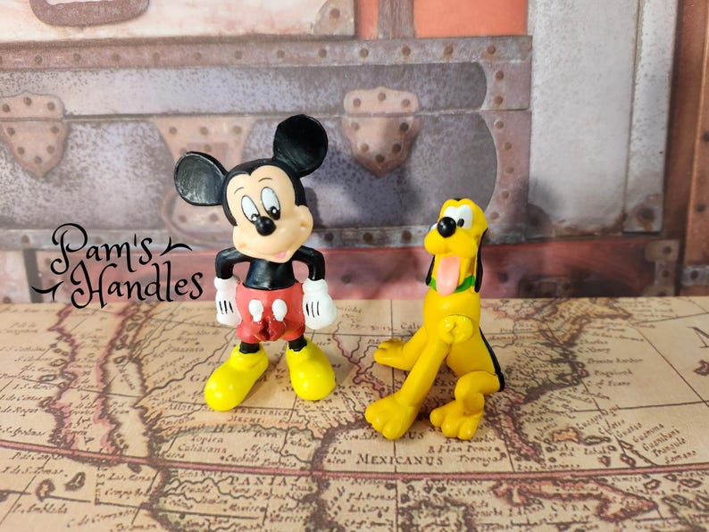 以下が含まれることがあります： ミッキーマウスとプルートの2つのディズニーフィギュア。ミッキーマウスは赤いショートパンツ、黄色の靴、白い手袋をしています。プルートは、黒い耳と舌が出ている黄色の犬です。フィギュアは地図の上にあります。