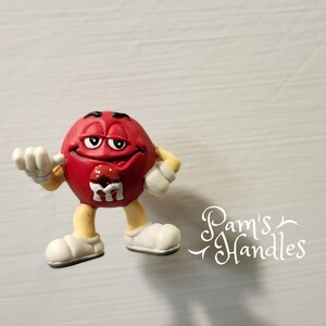 Peut inclure: Figurine rouge repr&eacute;sentant un personnage M&M's avec des gants et des chaussures blancs. La figurine a un visage souriant et un petit M sur la poitrine. Le texte "Pam's Handles" est dans le coin inf&eacute;rieur droit.