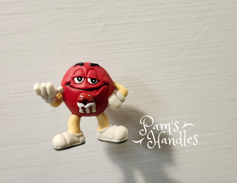 Peut inclure: Figurine rouge repr&eacute;sentant un personnage M&M's avec des gants et des chaussures blancs. La figurine a un visage souriant et un petit M sur la poitrine. Le texte "Pam's Handles" est dans le coin inf&eacute;rieur droit.