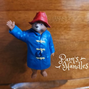 May include: A figurine of Paddington Bear, wearing a blue coat and a red hat, waving with his right paw. The bear has a brown face and is standing on a wooden surface. The figurine is a decorative item.