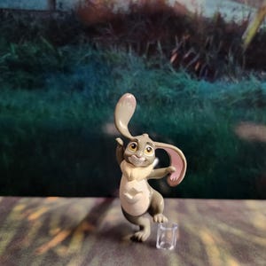Puede incluir: Una figurita de conejo gris y blanco con orejas rosas, de pie sobre una pierna con la otra pierna doblada. La figurita lleva un collar de piel blanca y tiene un diseño en forma de estrella en el pecho. La figurita está de pie sobre una base de plástico transparente.