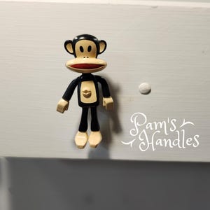 Op de afbeelding: Een Paul Frank apenladeknop in zwart, bruin en rood, gemonteerd op een witte lade. De apenvormige handgreep heeft een zwarte body, bruine handen en voeten en een rode mond. De tekst "Pam's Handles" is cursief geschreven.