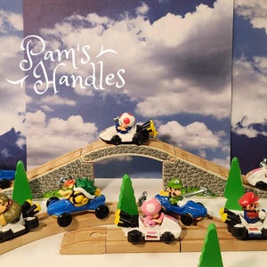 Pode incluir: Uma coleção de figuras de personagens de Mario Kart em carros de corrida em miniatura, incluindo Mario, Luigi, Toad, Peach, Yoshi, Donkey Kong e Bowser, dispostos em pistas de madeira com uma ponte de pedra e árvores de plástico verde.