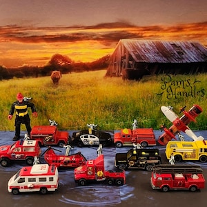 Pode incluir: Uma coleção de caminhões de bombeiros de brinquedo em miniatura, um avião e uma figura de bombeiro. Os brinquedos são dispostos em uma superfície que parece um rio com um celeiro de madeira ao fundo. Os brinquedos são todos vermelhos e amarelos com detalhes pretos. A figura do bombeiro está usando um capacete vermelho e um uniforme preto. Os brinquedos estão todos em diferentes estágios de montagem. O texto "Pam's Handles" é visível no fundo.