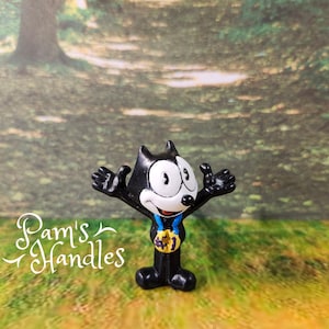 May include: A black and white Felix the Cat figurine with arms outstretched, wearing a blue ribbon and a gold medal. The figurine is standing on a green surface with a blurred background of trees. The text "Pam's Handles" is in the image.