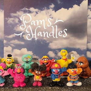 May include: A collection of colourful Sesame Street character figurines including Elmo, Cookie Monster, Big Bird, and others, arranged in front of a blue sky backdrop with the white text Pam's Handles printed in the centre of the image.