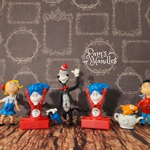 Könnte beinhalten: Eine Sammlung von Dr. Seuss-Charakterfiguren, darunter der Kater mit Hut, die Stempel Thing 1 und Thing 2, Sally und ein Junge mit einem Fisch in einer Teekanne. Die Figuren sind vor einem gemusterten Hintergrund platziert.