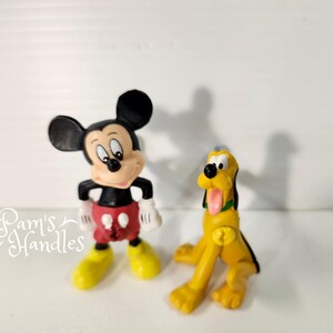 以下が含まれることがあります： 2つのディズニーフィギュア：ミッキーマウスとプルート。ミッキーマウスはクラシックなポーズで、黒いショートパンツ、赤いショートパンツ、黄色の靴を履いています。プルートは黄色で、黒い耳とピンクの舌をしています。
