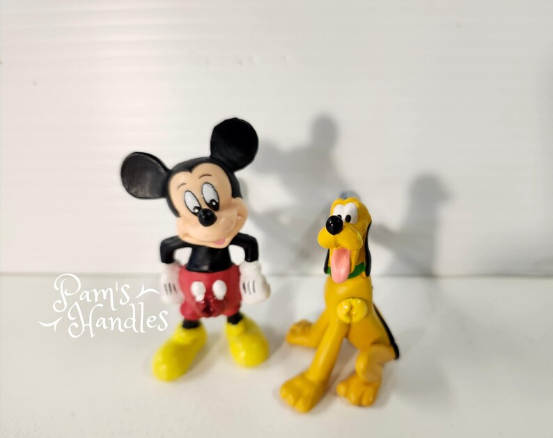 以下が含まれることがあります： 2つのディズニーフィギュア：ミッキーマウスとプルート。ミッキーマウスはクラシックなポーズで、黒いショートパンツ、赤いショートパンツ、黄色の靴を履いています。プルートは黄色で、黒い耳とピンクの舌をしています。