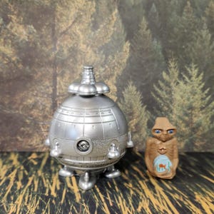Op de afbeelding: Een zilveren speelgoed ruimteschip met een koepelvormige bovenkant en drie poten, naast een bruin alien figuurtje met grote ogen.