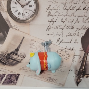 Op de afbeelding: Een blauwe en rode opwindbaar speelgoed olifant met een gele kroon. Het speelgoed ligt op een vintage achtergrond met een klok, een ansichtkaart van de Eiffeltoren en een pen.