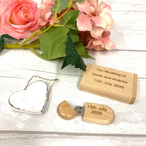 May include: A wooden USB drive shaped like a heart with the text "13th July 2020" engraved on it. The drive is open, revealing the USB connector. A small wooden box with the text "The Wedding of Sarah and Andrew 13th July 2020" engraved on it is also shown. A white heart-shaped ornament and pink flowers are in the background.