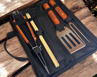 May include: A black zippered carrying case containing a set of grilling tools with wooden handles. The set includes metal tongs, a set of wooden skewers, a slotted spatula, and a two-pronged fork. Each tool handle is engraved with the text SAM.