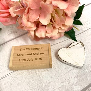 May include: A small wooden box with the text "The Wedding of Sarah and Andrew 13th July 2020" engraved on the top. The box is sitting on a white wooden surface with a white heart-shaped ornament next to it.