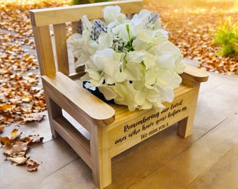 May include: A small wooden bench with a white flower arrangement on top. The bench has a personalized inscription that reads "Remembering our loved ones who have gone before us. We miss you x".