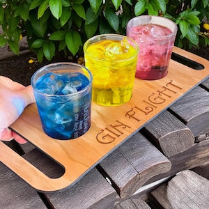 May include: A wooden serving tray with three cocktails in blue, yellow, and pink. Each drink is filled with ice. The tray is engraved with "GIN FLIGHT" and has a handle. The blue drink has text that reads "Autumn 15th Sept 2022".